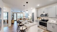 This thoughtfully designed single-floor residence offers a cohesive open-concept layout that seamlessly connects the kitchen, dining, and living areas. The kitchen is a standout feature, showcasing gray shaker-style cabinetry, a subway tile backsplash, stainless steel appliances including a refrigerator, range, and microwave, and a granite-topped island with an undermount sink and bar seating — ideal for casual dining and entertaining. Stylish globe pendant lights hang above the island, adding a contemporary touch.

The living room is bright and welcoming, benefiting from natural light through large windows and a glass door that opens to the outdoors. A ceiling fan overhead and wood-look plank flooring throughout the main living areas contribute to a clean, modern aesthetic.

The bedroom offers a comfortable, carpeted retreat with ample wall space and access to the dedicated closet. The bathroom is well-appointed with a vanity featuring blue-gray cabinetry, granite countertops, wood-look flooring, and a tub-shower combination with marble-style tile surround. The layout ensures convenient access between the bathroom and bedroom, creating a functional and cohesive private living zone.