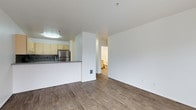 This single-floor unit is designed with an open-concept layout that connects the living area and kitchen, creating a functional and cohesive space. The flooring throughout is a warm-toned wood-look hardwood, lending a clean and contemporary feel to the interior.

The kitchen is partially separated from the living area by a raised breakfast bar with a dark countertop, offering a casual dining or prep surface. Light yellow cabinetry lines the kitchen walls, complemented by a tile backsplash and a stainless steel refrigerator that adds a modern touch. A flush-mount ceiling light illuminates the kitchen area effectively.

The living space features crisp white walls and a structural column that subtly defines zones within the open floor plan. A hallway visible from the main living area leads to a bathroom, which is partially visible and features light-colored fixtures and walls.

The unit is well-lit with a combination of ambient ceiling fixtures and natural light. The overall layout is practical and straightforward, with smooth wall finishes and consistent flooring tying the spaces together for a cohesive, move-in ready aesthetic.