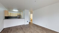 This single-floor unit is designed with an open-concept layout that connects the living area and kitchen, creating a functional and cohesive space. The flooring throughout is a warm-toned wood-look hardwood, lending a clean and contemporary feel to the interior.

The kitchen is partially separated from the living area by a raised breakfast bar with a dark countertop, offering a casual dining or prep surface. Light yellow cabinetry lines the kitchen walls, complemented by a tile backsplash and a stainless steel refrigerator that adds a modern touch. A flush-mount ceiling light illuminates the kitchen area effectively.

The living space features crisp white walls and a structural column that subtly defines zones within the open floor plan. A hallway visible from the main living area leads to a bathroom, which is partially visible and features light-colored fixtures and walls.

The unit is well-lit with a combination of ambient ceiling fixtures and natural light. The overall layout is practical and straightforward, with smooth wall finishes and consistent flooring tying the spaces together for a cohesive, move-in ready aesthetic.