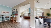 Located at 200 Blakeslee St in Bristol, Connecticut, this single-story community clubhouse offers a bright and inviting atmosphere designed for social gathering and relaxation. The open-plan layout is defined by elegant arched doorways and white structural columns that create natural zones while maintaining a sense of spaciousness throughout.

The main gathering area commands attention with soaring vaulted ceilings and dramatic arched windows that flood the space with natural light and offer views of the surrounding greenery. A cozy seating arrangement with leather armchairs anchors this sunlit lounge, complemented by a fireplace and a mounted television. Durable wood-look flooring runs throughout, tying the various zones together seamlessly.

A dedicated kitchen area features cream cabinetry, stainless steel appliances including a full-size refrigerator, and a peninsula counter with bar stool seating — ideal for casual dining or socializing. Adjacent to the kitchen, open shelving and built-in cabinetry provide additional display and storage space.

Multiple bistro-height tables with modern upholstered chairs are strategically placed throughout the space, offering flexible seating options for individuals or small groups. Soft, neutral wall tones accented by muted blue feature walls contribute to the calm and welcoming ambiance. Overall, the space is thoughtfully designed to foster community connection in a comfortable, well-lit environment.