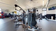 This fitness center is a thoughtfully designed single-floor facility that offers a wide range of workout options within a spacious, open layout. The interior features high ceilings with exposed ductwork and large ceiling fans, contributing to an industrial-modern aesthetic. Linear LED lighting runs throughout, ensuring the space is well-lit at all hours.

Large windows line multiple walls, allowing abundant natural light to flood the interior and providing views of the surrounding outdoor environment. The upper clerestory windows further enhance the brightness and sense of openness. A bold "WORK IT" graphic on the wall adds a motivating, energetic touch to the atmosphere.

Durable rubber flooring covers the main workout areas, with green turf matting designated for free weight and functional training zones. The facility is equipped with a full array of commercial-grade strength machines, including cable systems and resistance equipment, alongside a dedicated free weight area with a full dumbbell rack and adjustable benches. Cardio equipment includes treadmills, ellipticals, stair climbers, and rowing machines. A functional training zone features a TRX station and a heavy tire for dynamic exercises. Wall-mounted televisions provide entertainment throughout.