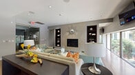 This elegantly designed single-floor communal space blends modern architecture with refined interior details, creating an inviting and functional environment. The centerpiece of the main lounge area is a floor-to-ceiling textured stone fireplace wall, adorned with a decorative sunburst mirror and flanked by dark wood built-in shelving niches. Comfortable sectional and accent seating, layered over plush area rugs and warm hardwood flooring, create a welcoming gathering atmosphere. Recessed lighting throughout ensures a well-lit, polished ambiance. A continuous band of large, low-profile windows runs along one wall, filling the lounge with natural light and offering views of the lush greenery outside. A wall-mounted flat-screen television adds a practical entertainment feature to the space. The gourmet kitchen is equipped with a professional-grade gas range, an expansive granite-topped island with dark wood cabinetry, stainless steel appliances including a full-size refrigerator, and a built-in window bench overlooking the outdoors. The transition areas feature glass sliding doors, additional built-in shelving, bamboo hardwood flooring, and curated artwork, tying the space together with a cohesive, contemporary design sensibility.