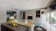 This elegantly designed single-floor communal space blends modern architecture with refined interior details, creating an inviting and functional environment. The centerpiece of the main lounge area is a floor-to-ceiling textured stone fireplace wall, adorned with a decorative sunburst mirror and flanked by dark wood built-in shelving niches. Comfortable sectional and accent seating, layered over plush area rugs and warm hardwood flooring, create a welcoming gathering atmosphere. Recessed lighting throughout ensures a well-lit, polished ambiance. A continuous band of large, low-profile windows runs along one wall, filling the lounge with natural light and offering views of the lush greenery outside. A wall-mounted flat-screen television adds a practical entertainment feature to the space. The gourmet kitchen is equipped with a professional-grade gas range, an expansive granite-topped island with dark wood cabinetry, stainless steel appliances including a full-size refrigerator, and a built-in window bench overlooking the outdoors. The transition areas feature glass sliding doors, additional built-in shelving, bamboo hardwood flooring, and curated artwork, tying the space together with a cohesive, contemporary design sensibility.