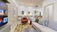 Located at 902 Lytton St in North Vancouver, BC, this thoughtfully designed single-floor amenity space offers a welcoming and contemporary environment. The layout encompasses a living room and a dining room, together creating a cohesive and functional communal area finished with light hardwood flooring throughout.

The living room is a bright and inviting retreat, bathed in natural light from large floor-to-ceiling windows that frame verdant views of surrounding trees. A slatted wood accent wall anchors a wall-mounted flat-screen television, while curated furnishings — including a plush sectional sofa, a terracotta accent chair, and a patterned area rug — add warmth and character. A built-in counter with bar seating and ambient lighting provides a versatile workspace or casual lounge area, complemented by contemporary artwork on the walls.

The dining room is equally well-appointed, featuring a long dark-topped communal table surrounded by natural wood chairs, ideal for group gatherings. A kitchenette alcove with light wood cabinetry, white tile backsplash, a stainless steel refrigerator, and a sink supports the space's functionality. Recessed lighting and direct exterior access via a glass-paneled door further enhance the airy, open atmosphere of this well-curated space.