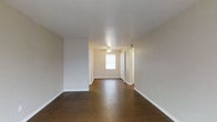 This single-floor space presents a clean, open living area designed with simplicity and functionality in mind. The room is finished with rich, dark hardwood flooring that extends throughout, providing a warm contrast against the light, neutral-toned walls. A textured white ceiling adds subtle depth overhead, while a ceiling fan with a built-in light fixture serves as the central overhead lighting source, offering both comfort and practicality.

Natural light enters through a window positioned near what appears to be the entryway, brightening the space and creating an inviting atmosphere. The entryway area features white doors — likely a closet and the main entry — framed by clean white trim that complements the overall neutral palette. White baseboards run along the perimeter of the room, adding a crisp, finished look to the space.

The open layout provides a generous sense of space, making it versatile for various furniture arrangements. The absence of clutter and the minimal design aesthetic give the room a fresh, move-in-ready feel. Overall, the space offers a straightforward, well-maintained environment with consistent finishes throughout.