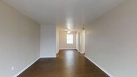 This single-floor space presents a clean, open living area designed with simplicity and functionality in mind. The room is finished with rich, dark hardwood flooring that extends throughout, providing a warm contrast against the light, neutral-toned walls. A textured white ceiling adds subtle depth overhead, while a ceiling fan with a built-in light fixture serves as the central overhead lighting source, offering both comfort and practicality.

Natural light enters through a window positioned near what appears to be the entryway, brightening the space and creating an inviting atmosphere. The entryway area features white doors — likely a closet and the main entry — framed by clean white trim that complements the overall neutral palette. White baseboards run along the perimeter of the room, adding a crisp, finished look to the space.

The open layout provides a generous sense of space, making it versatile for various furniture arrangements. The absence of clutter and the minimal design aesthetic give the room a fresh, move-in-ready feel. Overall, the space offers a straightforward, well-maintained environment with consistent finishes throughout.