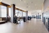 Located at 3201 N Federal Hwy in Oakland Park, Florida, this contemporary fitness center offers a generously sized, single-floor layout designed to accommodate a wide range of workout styles and training needs. The space is defined by its sleek, modern aesthetic, characterized by a neutral palette of whites, grays, and dark charcoal accents, complemented by warm linear lighting embedded in the ceilings.

The main workout floor is expansive and open, finished with durable rubber flooring and outfitted with a comprehensive selection of strength machines, cable systems, free weights, benches, and cardio equipment including treadmills and ellipticals. Floor-to-ceiling windows line the perimeter, flooding the space with natural light and offering impressive elevated views of the surrounding South Florida skyline and sky.

A dedicated studio area features light hardwood flooring, full-wall mirrors, recessed cove lighting, and ample open floor space suitable for yoga, group fitness, or personal training sessions. An accessory zone provides stability balls, medicine balls, resistance bands, and functional training tools.

The entry area showcases a tall slatted wood accent wall, adding warmth and texture to the otherwise contemporary design. Throughout, large glass partitions maintain visual openness while defining distinct training zones, creating a professional yet inviting fitness environment.