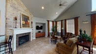 Located at 11655 Briar Forest Dr in Houston, TX 77077, this single-floor community space offers an open and welcoming layout defined by soaring vaulted ceilings and generous natural light throughout. The main living area serves as the centerpiece, showcasing a striking floor-to-ceiling natural stone fireplace surround with a clean white mantle and a built-in media niche equipped with cabinetry below. Recessed lighting and a ceiling fan complement the vaulted ceiling, while large windows dressed with rich, earth-toned drapery panels and wood blinds frame the room and allow abundant daylight to filter in. Tile flooring in a warm neutral tone extends throughout the space, providing a cohesive and easy-to-maintain surface. An adjacent dining zone features high-top bistro tables and bar-height seating, opening through a wide doorway to an additional windowed room, ideal for smaller gatherings. The space transitions into a spacious gathering hall with dramatically high ceilings anchored by a grand arched window, flooding the area with light. A pass-through bar counter with pendant lighting and granite-topped surfaces connects to a service area, making this layout well-suited for social events and community functions. The overall ambiance is warm, inviting, and purposefully designed for shared use.