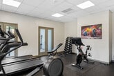 This fitness center is a thoughtfully designed, single-room facility that maximizes its compact footprint to deliver a comprehensive workout environment. The space features durable speckled rubber flooring throughout, providing both safety and resilience underfoot, while a suspended drop-tile ceiling with recessed panel lighting ensures consistent, bright illumination across the entire room.

The cardio zone is well-appointed, housing treadmills, an elliptical trainer, a rowing machine, and a Peloton stationary bike with an integrated display screen. A dumbbell rack offers a range of free weights for strength training, complemented by a padded adjustable workout bench and a BOSU balance trainer for functional fitness. A wall-mounted television adds entertainment options during workouts.

Natural light flows in through multiple large windows and glass-panel doors positioned along two walls, creating an open, airy atmosphere. The doors provide direct outdoor access to what appears to be a pool area, seamlessly connecting indoor fitness with outdoor amenities. A water dispenser is conveniently available within the space.

The white walls are accented by vibrant, colorful fitness-themed artwork depicting dumbbells and kettlebells, adding an energetic visual character to the room. Wooden shelving units provide practical storage. Overall, the fitness center presents a clean, functional, and motivating environment suited for a well-rounded daily workout routine.