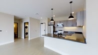 This contemporary single-floor residence showcases a thoughtfully designed open-concept layout that seamlessly blends living and kitchen spaces. The kitchen is a standout feature, equipped with stainless steel appliances including a French door refrigerator, electric range, and built-in microwave. Two-tone cabinetry in a warm wood-grain finish provides ample storage, while the expansive kitchen island with a dark granite countertop offers both additional workspace and a natural gathering point. A built-in sink with a sleek matte black faucet adds a refined touch to the island's design. Overhead, a pair of black industrial-style pendant lights illuminate the space, contributing to the modern aesthetic. Light wood-toned flooring runs throughout, complementing the clean white walls and creating a bright, airy atmosphere. Recessed lighting in the ceiling enhances the sense of openness. A dedicated in-unit laundry closet discreetly houses a stacked washer and dryer, offering everyday convenience. The overall design is clean, functional, and modern, making this a well-appointed space suited for contemporary living.