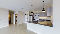 This contemporary single-floor residence showcases a thoughtfully designed open-concept layout that seamlessly blends living and kitchen spaces. The kitchen is a standout feature, equipped with stainless steel appliances including a French door refrigerator, electric range, and built-in microwave. Two-tone cabinetry in a warm wood-grain finish provides ample storage, while the expansive kitchen island with a dark granite countertop offers both additional workspace and a natural gathering point. A built-in sink with a sleek matte black faucet adds a refined touch to the island's design. Overhead, a pair of black industrial-style pendant lights illuminate the space, contributing to the modern aesthetic. Light wood-toned flooring runs throughout, complementing the clean white walls and creating a bright, airy atmosphere. Recessed lighting in the ceiling enhances the sense of openness. A dedicated in-unit laundry closet discreetly houses a stacked washer and dryer, offering everyday convenience. The overall design is clean, functional, and modern, making this a well-appointed space suited for contemporary living.
