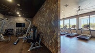 This fitness center is thoughtfully designed to support a full range of workout routines, divided into two distinct zones separated by a striking textured stone accent wall in a herringbone pattern. The cardio area is bright and inviting, lined with large floor-to-ceiling windows that flood the space with natural light and offer views of an outdoor pool and surrounding greenery. A row of Matrix treadmills faces the windows, complemented by elliptical machines and stair climbers along the adjacent wall. A bold motivational mural reading "The Stronger You Are, The Better You Feel" energizes the space, while a large ceiling fan and recessed lighting ensure comfort during workouts. The strength training zone features a darker, more focused atmosphere with a dark ceiling, rubber flooring, and spotlighting. It is equipped with multiple cable and resistance machines, a dumbbell rack, and a wall-mounted television. Full-length mirrors amplify the space and support proper form. A separate open area near the entrance provides additional open floor space for stretching or functional training, with sanitation stations conveniently placed throughout. The overall design balances industrial aesthetics with practical functionality.