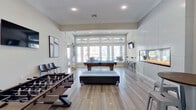 This stylish, single-floor game and lounge space is designed for social gathering and recreation, combining contemporary aesthetics with a range of entertainment amenities. The open layout is anchored by light wood-look tile flooring that runs throughout, lending a clean, cohesive look to the entire room.

At the heart of the space sits a full-size pool table with a vibrant blue felt surface, positioned beneath a generous bank of large windows and a glass door that frame views of an adjacent outdoor patio and pool area, filling the room with abundant natural light. A foosball table occupies one side of the room alongside a wall-mounted flat-screen television, flanked by monochromatic abstract artwork. Sleek bar-height chairs and a round bistro table provide casual seating nearby.

A striking floor-to-ceiling panel wall houses a wide, linear gas fireplace, serving as a dramatic focal point on one side of the room. Additional bar stools along a countertop ledge create an informal gathering spot. Sliding barn-style doors with frosted glass panels provide access to a separate adjoining area, adding a modern-industrial design detail. Recessed lighting and elevated ceilings contribute to the open, airy ambiance throughout.