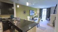 This single-floor residence showcases an open-concept design that seamlessly connects the kitchen, dining, and living areas into one cohesive living space. The kitchen is appointed with dark granite countertops and a functional island that doubles as a breakfast bar, complemented by pendant lighting that adds both style and task illumination. A stainless steel sink and modern cabinetry complete the kitchen's polished look.

The dining area sits just beyond the kitchen island, featuring a glass-top table with upholstered chairs, positioned beneath additional pendant and track lighting. A bold green accent wall serves as a striking visual anchor throughout the open layout, adorned with decorative wall art and a starburst mirror that adds texture and dimension.

The living area flows naturally from the dining space, featuring plush carpeting, a comfortable sofa, and coordinating accent pieces. Bold patterned drapes frame a large window that allows abundant natural light to fill the space and offers views of a balcony beyond. The overall ambiance is modern, vibrant, and thoughtfully curated with cohesive color tones throughout.