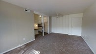 Located at 412 Thurber Dr W in Columbus, Ohio, this single-floor unit offers a straightforward and functional layout designed for comfortable everyday living. The main living area is generously proportioned, featuring wall-to-wall carpet flooring that adds warmth underfoot and neutral-toned walls that provide a clean, versatile backdrop for any furnishing style. Natural light filters into the space, further brightening the open feel of the room. A notable feature along one wall is a set of large built-in white panel storage units, offering substantial organizational capacity while maintaining a tidy, streamlined appearance. The entry door is conveniently positioned adjacent to this storage wall. The kitchen is accessible directly from the living area and is defined by a partial partition wall that maintains an open sightline between the two spaces. It is equipped with essential appliances including a refrigerator, and features counter space with cabinetry above. Open shelving is incorporated into the kitchen area, adding both display and storage functionality. The ceiling-mounted smoke detector and HVAC vent are visible throughout, reflecting attention to safety and climate control. Overall, the space presents a clean, move-in ready environment with practical features suited for urban Columbus living.