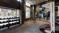 The 290 George St Fitness Center in New Brunswick, NJ is a thoughtfully designed, single-floor facility that provides a comprehensive workout environment for all fitness levels. The space features soaring ceilings with large street-facing windows that flood the gym with natural light and offer views of the surrounding neighborhood, creating an open and energizing atmosphere. Dark, sophisticated wall tones are complemented by a distinctive decorative accent wall with a vibrant teal and yellow geometric mural, adding a dynamic visual element to the space. The flooring throughout consists of durable, textured carpet tiles suited for high-traffic athletic use. The fitness center is equipped with a wide range of cardio machines, including treadmills, ellipticals, and stationary bikes, as well as a selection of selectorized strength training machines. A dedicated free weights area offers a full rack of dumbbells, kettlebells, and medicine balls. Functional training is supported by a TRX suspension system, a mini trampoline, and a freestanding Century punching bag. Wall-mounted flat-screen TVs are positioned throughout for entertainment during workouts. Recessed and track lighting ensures the entire space is well-lit. The facility is accessed through glass entry doors directly from George Street, making it conveniently accessible.