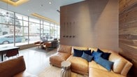 This contemporary clubhouse is thoughtfully designed to serve as a dynamic communal gathering space. Large floor-to-ceiling grid-pane windows dominate the exterior-facing walls, flooding the interior with natural light and offering open views of the surrounding streetscape. The lounge area features plush leather sectional sofas accented with rich navy blue throw pillows, complemented by warm cognac-toned accent chairs and sculptural side tables. A textured taupe feature wall with decorative wall hooks anchors the seating area, while cove lighting along the ceiling adds a warm, ambient glow. An intimate media nook is defined by a full wood-clad accent wall with natural grain detailing, a wall-mounted flat-screen television, and a cozy sectional seating arrangement atop a patterned area rug. The kitchen is a standout feature, showcasing a lengthy marble-topped island with integrated bar seating, white hexagonal mosaic tile flooring, full-height subway tile walls, stainless steel appliances, a deep sink, and open shelving stocked with books and décor. A dining area with dark wood tables and red chairs sits adjacent, completing the space. Together, these areas create a cohesive, upscale environment ideal for socializing and community engagement.