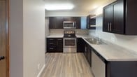 This single-floor, two-bedroom, one-bathroom unit offers a practical and well-appointed layout designed for comfortable everyday living. The open living area features dark wood-look vinyl flooring and a ceiling fan overhead, with a large picture window fitted with vertical blinds allowing generous natural light to fill the space. A baseboard heating system runs along the walls, providing efficient warmth throughout.

The kitchen is thoughtfully designed with dark espresso cabinetry, light countertops, and a full suite of stainless steel appliances, including a refrigerator, electric range, built-in microwave, and dishwasher. Light wood-look flooring and ample upper and lower cabinet storage make this a highly functional cooking space.

Both bedrooms are carpeted for comfort and each feature windows with blinds, baseboard heating, and neutral walls. The dual-window configuration in one bedroom provides additional light and ventilation.

The bathroom is well-equipped with dark vanity cabinetry, a white countertop with an integrated sink, a large mirror with overhead lighting, tiled flooring, and a full tub-shower combination. The unit's consistent use of warm wood tones and neutral finishes throughout creates a cohesive and inviting atmosphere.