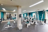 This fitness center offers a generously sized, single-floor workout facility designed to accommodate a wide range of exercise needs. The space is defined by bold teal accent walls contrasted against neutral beige tones, creating an energizing yet clean aesthetic throughout. Durable speckled rubber flooring covers the main workout areas, providing both safety and comfort underfoot.

The strength training zone is well-appointed with a comprehensive array of white-framed cable and resistance machines, featuring teal-upholstered seats and pads. A full-length mirrored wall runs along one side of the room, visually expanding the space and allowing users to monitor their form during exercise. A dedicated free weights area includes a rack of dumbbells and a selection of medicine balls and stability equipment.

A separate cardio alcove houses three treadmills positioned in front of a wall-mounted flat-screen television, offering an entertainment option for cardio sessions. A coat rack and bench provide practical convenience in this area.

Recessed and flush-mounted ceiling light fixtures ensure consistent, even illumination throughout the facility. Large windows and a glass-paned exterior door allow natural light to filter in, providing views of the surrounding greenery. The layout is open and uncluttered, with structural columns integrated seamlessly into the floor plan, making for an efficient and functional workout environment.