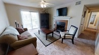 The living area of this single-floor space is designed for comfort and functionality. Plush wall-to-wall carpeting anchors the room, providing a warm foundation beneath a patterned area rug that defines the main seating area. A corner fireplace serves as a natural focal point, framed by a classic white mantel and tile surround, with a wall-mounted flat-screen television positioned above for a streamlined entertainment setup. A ceiling fan with integrated lighting occupies the center of the room, contributing to both air circulation and ambient illumination. Natural light flows generously into the space through a large sliding glass door fitted with vertical blinds, which opens to an outdoor balcony or patio area, extending the living space and offering views of the surrounding environment. A dark wood armoire adds storage and visual contrast against the neutral wall tones. The overall ambiance is warm and inviting, with a traditional aesthetic conveyed through the combination of rich wood furnishings, neutral carpeting, and classic architectural details. The layout is practical and well-proportioned, making efficient use of the available floor space.
