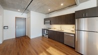 This single-floor urban loft showcases an industrial-modern aesthetic that blends raw architectural elements with contemporary finishes. The open-plan layout is anchored by exposed concrete ceilings and visible ductwork, lending an authentic loft character throughout the space. Warm hardwood flooring flows across the main living and kitchen areas, creating a striking contrast against the cool concrete overhead.

The kitchen is thoughtfully designed with dark espresso cabinetry, stainless steel appliances including a range, microwave, and dishwasher, and a classic white subway tile backsplash that adds a clean, timeless touch. Recessed lighting illuminates the workspace efficiently.

A sliding concrete-panel barn door serves as a functional room divider, adding to the industrial character of the space. Large, multi-pane windows flood the interior with natural light and frame panoramic views of a lush, tree-lined hillside, creating a bright and airy atmosphere.

The bathroom features a clean, minimalist design with a dark wood vanity, backlit mirror, white countertop, and a walk-in shower with a curtain enclosure. Neutral flooring and crisp white walls maintain the space's modern sensibility. Overall, this loft offers a cohesive blend of industrial character and modern comfort.