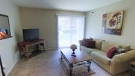 This single-floor space centers around a comfortable living room finished with wall-to-wall carpeting in a neutral tone, contributing to a warm and inviting atmosphere. The room is anchored by a large sliding glass door dressed with vertical blinds, allowing abundant natural light to filter in while providing a view of the greenery outside. The beige walls offer a clean, understated backdrop that complements the room's overall neutral palette. A traditional-style wooden coffee table with ornate metal legs rests atop a patterned area rug, serving as the focal point of the seating arrangement. The space accommodates a generously sized sofa adorned with decorative pillows. Along one wall, a compact wooden desk with a lower shelf provides a functional area for a television or workspace setup. Ceiling-mounted air vents are visible, indicating a central HVAC system. The layout is open and uncluttered, maximizing usable floor space. Overall, the living area presents a practical yet comfortable environment with classic furnishing details and a modest, well-maintained aesthetic throughout.