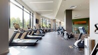 Located at 7970 Maitland St in McLean, Virginia, this contemporary fitness center is thoughtfully designed to provide a comprehensive and motivating workout environment. The single-floor facility is organized into distinct zones that cater to a wide range of fitness activities.

The main cardio area is a standout feature, lined with an extensive row of treadmills, ellipticals, and stationary bikes from Matrix. Floor-to-ceiling windows run along the length of this section, flooding the space with natural light and offering views of the surrounding greenery — creating an energizing atmosphere for cardio training. Wall-mounted televisions are positioned for convenient viewing during workouts.

The strength training zone offers a well-equipped selection of cable machines, weight racks, free weights, benches, and functional training equipment. This area benefits from both recessed and linear LED lighting, maintaining a bright and focused environment. A bold geometric accent wall in sage green adds a modern design touch to this section.

Throughout the facility, large-scale artistic wall murals depicting fitness-themed imagery — including bicycles, kettlebells, trophies, and stopwatches — lend a dynamic and energetic character to the space. The entryway features a striking full-wall mural that sets the tone upon arrival.

Durable speckled rubber flooring is used throughout, providing both safety and a polished, professional aesthetic.