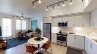 This single-floor unit showcases a contemporary open-concept layout that seamlessly connects the kitchen, dining, and living areas. The kitchen is outfitted with stainless steel appliances, including a full-size refrigerator, range, and over-the-range microwave, complemented by sleek white cabinetry, light countertops, and a classic white subway tile backsplash. Track lighting runs along the ceiling, providing focused illumination throughout the space. The dining area features a round white pedestal table paired with cognac-toned chairs, creating an inviting spot for meals. The living area extends naturally from the dining space, anchored by a ceiling fan, neutral furnishings, and colorful wall art that adds a lively touch. Light wood-style flooring runs throughout, enhancing the bright, airy feel of the interior. A private balcony extends from the unit and overlooks a resort-style pool area shaded by blue umbrellas, surrounded by tall palm trees and landscaped grounds. The stucco exterior and black metal railing frame sunny outdoor views, characteristic of a warm-climate community setting.