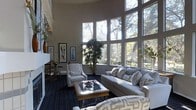 This refined single-floor residence makes a striking first impression with its dramatic, soaring ceilings and an expansive curved wall of floor-to-ceiling windows that floods the living area with natural light. The windows frame a lush, wooded outdoor backdrop, seamlessly blending the interior with nature and creating a tranquil, light-filled atmosphere throughout the day.

The living room serves as the centerpiece of the space, thoughtfully appointed with a plush, neutral-toned sectional sofa accented by patterned throw pillows in a classic geometric motif. Complementing the seating arrangement are matching patterned armchairs, lending a cohesive and polished aesthetic. A solid wood coffee table with a glass insert anchors the seating area, adding warmth and texture.

The deep-hued, patterned carpet grounds the room and contrasts elegantly with the warm beige walls and bright, airy windows. Artful touches — including framed wall art, decorative sculptural vases, and lush indoor plants — enrich the space with personality and visual interest. A fireplace adds an element of warmth and classic charm to the room's overall composition.

The interplay of natural light, thoughtful furnishings, and architectural details creates an inviting, sophisticated ambiance ideal for both relaxed living and refined entertaining.