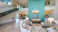 Located at 9200 Bruckhaus St in Raleigh, NC, this beautifully appointed community clubhouse offers an upscale, resort-inspired atmosphere defined by a cohesive teal and aqua color palette. The centerpiece of the space is a dramatic, floor-to-ceiling turquoise mosaic tile accent wall that anchors the main lounge area, drawing the eye upward and emphasizing the impressive double-height ceilings. A sleek floating staircase with glass panel railings and wood-toned treads adds an architectural focal point while connecting to an upper mezzanine level overlooking the main floor. Large floor-to-ceiling windows invite abundant natural light and provide views of the surrounding exterior. The main lounge features multiple sophisticated seating arrangements with neutral upholstered sofas and chairs accented by teal throw pillows, glass-top side tables, and carefully curated contemporary artwork. A secondary lounge area includes vibrant teal accent chairs arranged around a modern fireplace with stacked stone surround and a wall-mounted flat-screen TV, complemented by a textured gold honeycomb wall panel. Large-format stone tile flooring flows seamlessly throughout, unifying the open-plan layout. A kitchenette area provides dark wood cabinetry, quartz countertops, and a metallic tile backsplash. Suspended circular pendant lighting adds warmth and a polished finishing touch to this thoughtfully designed communal space.