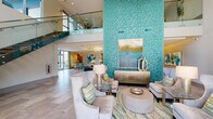 Located at 9200 Bruckhaus St in Raleigh, NC, this beautifully appointed community clubhouse offers an upscale, resort-inspired atmosphere defined by a cohesive teal and aqua color palette. The centerpiece of the space is a dramatic, floor-to-ceiling turquoise mosaic tile accent wall that anchors the main lounge area, drawing the eye upward and emphasizing the impressive double-height ceilings. A sleek floating staircase with glass panel railings and wood-toned treads adds an architectural focal point while connecting to an upper mezzanine level overlooking the main floor. Large floor-to-ceiling windows invite abundant natural light and provide views of the surrounding exterior. The main lounge features multiple sophisticated seating arrangements with neutral upholstered sofas and chairs accented by teal throw pillows, glass-top side tables, and carefully curated contemporary artwork. A secondary lounge area includes vibrant teal accent chairs arranged around a modern fireplace with stacked stone surround and a wall-mounted flat-screen TV, complemented by a textured gold honeycomb wall panel. Large-format stone tile flooring flows seamlessly throughout, unifying the open-plan layout. A kitchenette area provides dark wood cabinetry, quartz countertops, and a metallic tile backsplash. Suspended circular pendant lighting adds warmth and a polished finishing touch to this thoughtfully designed communal space.