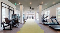 Located at 1701 J N Pease Pl in Charlotte, NC, this fitness center is a thoughtfully designed, single-floor facility offering a comprehensive workout environment. The space is defined by its open layout, supported by white structural columns that divide the floor into functional zones without compromising the sense of spaciousness.

The flooring features a distinctive geometric pattern combining yellow-green and gray carpet sections, adding visual interest throughout. Recessed lighting and large ceiling fans maintain a bright, comfortable atmosphere, while expansive floor-to-ceiling windows and glass doors along multiple walls allow abundant natural light and views of the surrounding outdoor landscape.

Cardio equipment is well-represented, with multiple treadmills, elliptical trainers, a stair climber, and stationary bikes distributed across the space. Strength training options include cable machines, a weight bench, and free weights. An interactive boxing-style trainer adds a dynamic fitness option. Full-length mirrors on one wall enhance the sense of depth and support form-checking during workouts.

A dedicated treadmill alcove features a dark-painted tray ceiling, recessed lighting, and windows overlooking greenery, creating a focused training sub-space. Glass interior doors lead to additional workout areas, and exterior doors provide direct outdoor access. The overall ambiance is modern, clean, and energizing.