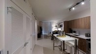 This single-floor residence showcases a thoughtfully designed open-concept layout that connects the kitchen and living areas in a cohesive, modern aesthetic. The interior is finished in a predominantly white palette, creating a clean and airy atmosphere throughout the space.

The kitchen area features warm wood cabinetry that provides a striking contrast against the bright surroundings. A sleek white high-top dining table with chrome-accented bar stools serves as a functional divider between the kitchen and living space, adding both style and practicality.

The living area is anchored by a patterned area rug and furnished with a contemporary sofa accented with mustard yellow throw pillows, complemented by a wall-mounted flat-screen television. Floor-to-ceiling sheer curtains line the far wall, diffusing natural light softly into the room and enhancing the open, welcoming ambiance.

Track lighting on the ceiling provides targeted illumination, while louvered bifold doors on the left side of the entryway add a classic architectural detail. A potted tropical plant near the window introduces a natural element, rounding out the modern yet inviting character of the space.