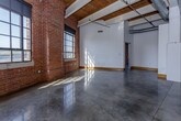 This single-floor urban loft showcases a compelling blend of industrial and modern design elements throughout. The open living area immediately draws attention with its soaring ceilings supported by exposed wood beams, polished concrete floors that reflect natural light, and prominent exposed brick walls that lend warmth and character to the industrial aesthetic. Large, multi-pane steel-framed windows flood the interior with abundant natural light while offering sweeping views of the surrounding cityscape.

The kitchen is thoughtfully appointed with warm wood cabinetry, frosted glass upper cabinets, dark granite countertops, a stainless steel farmhouse sink, and black appliances — all set beneath the dramatic open ceiling. A teal accent wall adds a vibrant pop of color to the open-plan living and kitchen area.

The bedroom features the same polished concrete flooring, exposed beam ceilings, and a mirrored sliding-door closet, maintaining the loft's cohesive design language. Two bathrooms complete the space: one with a sleek glass-top vanity, dark cabinetry, and exposed brick detail, and another with a slate-tiled walk-in shower, concrete floors, and light wood doors. Together, these elements create a sophisticated, industrial-chic living environment with a distinctly urban character.