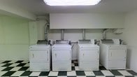 This single-floor laundry room is designed for functionality and efficiency. The space houses a row of top-load washers paired with front-load dryers, all finished in white and arranged along the back wall. Exposed ventilation ducts extend from the dryers upward, ensuring proper exhaust management. The room features a classic black-and-white checkered tile floor, adding a timeless visual contrast to the utilitarian setting. Overhead, fluorescent lighting provides bright, even illumination throughout the space. The walls are finished in a clean white, contributing to an organized and no-frills atmosphere. A built-in overhead cabinet or soffit runs along the upper back wall, lending a structured appearance to the layout. The room is clearly designed with practicality in mind, offering adequate space between units for ease of use. Overall, this laundry room provides a straightforward, well-equipped environment suited for regular, high-volume use.