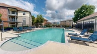 Located in Baton Rouge, Louisiana, this outdoor pool amenity area offers a well-appointed recreational retreat designed for relaxation and socializing. The centerpiece is a generously sized swimming pool with clear, blue water, surrounded by a broad pool deck finished in light-toned pavers. Numerous blue lounge chairs are arranged along the deck, providing ample space for sunbathing and leisure under the Louisiana sky.

Adjacent to the pool is a covered outdoor patio area that serves as an ideal gathering space. Outfitted with multiple dining tables and chairs, ceiling fans, and recessed lighting, this shaded zone ensures comfort even on warm days. Large glass sliding doors and windows connect the covered patio to the interior, allowing natural light to flow freely between spaces.

The entire pool area is enclosed by a sleek black metal security fence, with a gated entrance providing controlled access. Lush mature trees and manicured landscaping frame the perimeter, adding a touch of greenery and privacy. A separate pergola-style structure on the far end of the deck offers an additional shaded seating area. Views of the surrounding Baton Rouge skyline are visible beyond the fence, lending an urban backdrop to this inviting outdoor amenity space.