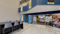 The Haven's lobby is a striking, open-concept entrance space defined by its soaring double-height ceilings and bold two-tone color scheme of deep navy blue and crisp white. The expansive tiled flooring features a decorative inlay pattern that anchors the central circulation area, contributing to the sense of openness and scale throughout the space.

A staffed reception desk sits prominently near the entrance, flanked by digital displays and branded "Welcome to The Haven" signage, establishing a professional and organized point of arrival. A branded pull-up banner further reinforces the identity of the space near the main entry doors, which allow natural light to filter into the interior.

Along one wall, a casual seating area with high-top tables and bar-height chairs provides a functional space for informal gatherings or brief meetings. Framed promotional graphics and imagery are displayed both at ground level and on the upper mezzanine-level walls, adding visual interest and drawing the eye upward.

Recessed lighting throughout the drop ceiling ensures consistent, even illumination across the lobby. The overall atmosphere is clean, modern, and welcoming, reflecting a well-organized and professionally maintained facility.