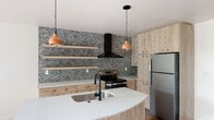 Located at 156 E 900 S in Salt Lake City, Utah, this single-floor space showcases a thoughtfully designed interior with contemporary finishes and a cohesive aesthetic. The kitchen serves as the centerpiece, anchored by a striking full-wall terrazzo backsplash that adds bold texture and visual depth. Light wood cabinetry with circular copper-toned pulls lines the walls, complemented by open floating shelves that provide both display and storage functionality. A curved kitchen island with a white quartz countertop and an undermount sink fitted with a matte black faucet offers a sleek, functional workspace. Overhead, warm terracotta pendant lights add an earthy contrast to the modern design. Stainless steel appliances, including a refrigerator and electric range with a black hood vent, complete the kitchen. Light wood-tone flooring flows throughout, reinforcing the warm, natural palette. The bathroom continues the design language with warm terracotta-toned wall tiles, a light wood vanity with quartz countertop, a matte black faucet, and an arched black-framed mirror. A soaking tub and dark slate-style floor tiles add to the bathroom's refined character. Outdoor views reveal lush, mature tree canopies and a glimpse of the surrounding mountains, bringing natural scenery into everyday living.