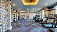 This expansive fitness center offers a generously sized, single-level layout designed to accommodate a full range of workout activities. The space is finished with a distinctive herringbone-patterned carpet throughout the main exercise area, transitioning to hardwood flooring in a dedicated functional training zone. Coffered ceilings with warm ambient lighting add architectural interest and an elevated ambiance to the room.

A row of modern cardio machines, including treadmills and ellipticals, lines the perimeter near floor-to-ceiling windows that invite abundant natural light and outdoor views. A Matrix functional trainer and a dedicated free weights area with a full dumbbell rack are positioned alongside large wall mirrors, maximizing both usability and the sense of openness in the space.

The functional training zone is highlighted by dramatic, large-scale illustrated murals depicting fitness-themed imagery — bicycles, kettlebells, and motivational typography — lending the space an energetic and inspiring character. This area also houses FORME interactive smart mirror stations alongside a dumbbell rack, supporting technology-driven personal training.

Structural columns punctuate the main floor, contributing to the layout's definition. Flat-screen TVs are mounted on the walls, and ceiling fans provide additional comfort. The overall design balances high-end aesthetics with practical, performance-focused functionality.