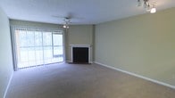 This single-floor space presents a straightforward and functional living area designed for everyday comfort. The main living room is generously sized, finished with wall-to-wall neutral-toned carpeting that adds warmth underfoot. A white-mantled fireplace serves as a focal point along one wall, providing both a decorative and functional element to the room. The walls are painted in a soft, neutral tone, complementing the overall understated aesthetic of the space.

Natural light enters through a large sliding glass door fitted with vertical blinds, which opens onto an outdoor balcony. The balcony provides views of a green, tree-lined exterior, bringing a sense of the outdoors into the living space. A ceiling fan with a built-in light fixture is centrally mounted, offering both air circulation and ambient lighting.

The textured popcorn ceiling is a notable architectural detail consistent throughout the space. White baseboards line the perimeter of the room, providing clean visual contrast against the carpeting and walls. Overall, the space presents a calm, neutral environment well-suited for comfortable day-to-day living.