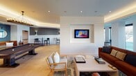 This single-floor communal space is thoughtfully designed to accommodate a variety of social activities, combining contemporary aesthetics with functional layout. The open floor plan is organized into several distinct zones that flow seamlessly into one another, unified by light herringbone-patterned hardwood flooring throughout.

The lounge area serves as the central hub, featuring two separate seating arrangements. One grouping centers around a shiplap-clad accent wall with a built-in flat-screen television, furnished with a light blue sofa accented by blue and white throw pillows. The second seating area offers a rich cognac leather sectional sofa positioned near floor-to-ceiling sliding glass doors that flood the space with natural light and provide outdoor views.

Adjacent to the main lounge, a billiards and entertainment zone features a full-sized pool table set beneath a statement chandelier, complemented by a dark accent wall adorned with round decorative mirrors. A casual high-top dining area with bistro-style tables and chairs sits along large windows overlooking the exterior.

The kitchenette is equipped with white cabinetry, a quartz countertop, stainless steel appliances including a microwave and beverage refrigerator, a utility sink, and a striking geometric-patterned tile backsplash illuminated by Edison-style pendant lights.