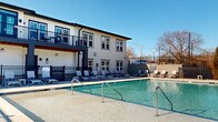 Located at 346 W 26th St in Charlotte, NC, this outdoor amenity space centers around a generously sized swimming pool with clear, blue-green water and gradual entry points equipped with stainless steel handrails. The pool area is designed with leisure and functionality in mind, offering a wide surrounding deck finished in light-toned concrete that provides ample space for relaxation and movement.

The sun deck is furnished with numerous lounge chairs and seating arrangements, accommodating a large number of guests simultaneously. The entire pool area is enclosed by a black iron fence, providing security while maintaining open sightlines to the surrounding landscape, which includes mature trees and views of neighboring residential buildings.

Adjacent to the pool, a modern two-story building features a covered ground-level terrace and an upper-level balcony with black metal railings and warm wood-toned ceiling accents. Large windows with black trim detail the exterior, which is finished in light siding complemented by bold dark architectural elements—reflecting a contemporary design aesthetic.

The outdoor setting benefits from abundant natural light throughout the day, with the open sky and surrounding greenery contributing to an inviting, resort-like atmosphere well-suited for outdoor recreation and social gatherings.