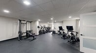 This single-floor fitness facility offers a thoughtfully designed wellness space organized across several distinct areas. The main workout floor is expansive, finished with durable dark rubber flooring and topped with a drop-panel ceiling equipped with recessed and flush lighting throughout. This primary area houses a cable weight machine with an adjustable bench, multiple treadmills, elliptical trainers, stationary cycling bikes with integrated screens, and a rack of dumbbells conveniently positioned near a mirrored window. The neutral white walls keep the space feeling clean and open.

Adjacent to the main floor are two dedicated yoga and stretching rooms finished with light wood-look vinyl flooring and warm greige walls. These calmer spaces feature natural light from windows dressed with sheer curtains, frosted-glass interior doors for privacy, and ample floor space for yoga mats and balance equipment such as BOSU balls. A small, neatly appointed towel station provides rolled towels on a marble-top side table, adding a welcoming touch. A restroom with wood-look flooring and clean white finishes rounds out the facility, ensuring convenience for all users.