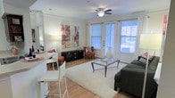 This single-floor residence offers a well-designed open layout that seamlessly connects the living and dining areas, creating an inviting and functional environment. The living room is a standout feature, bathed in natural light streaming through multiple large windows dressed with horizontal blinds and elegant draping. A ceiling fan with integrated lighting adds both comfort and a classic touch to the space.

The flooring throughout is warm hardwood, lending a clean, cohesive aesthetic that ties the entire space together. A plush white area rug anchors the living room seating area, which is centered around a sleek glass-top coffee table and complemented by a charcoal sofa and accent chairs in rich terracotta tones, creating a warm, inviting color palette.

A dark media console provides functional storage along one wall, while contemporary abstract artwork adds a vibrant pop of color. The dining area features a modern white bar-height counter with matching white bar stools, offering a casual and convenient spot for meals or entertaining.

Crown molding details along the ceiling add an architectural refinement to the space, while the overall open flow and abundant natural light create a bright, airy ambiance throughout this thoughtfully appointed single-floor home.