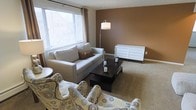 This single-floor space features a well-appointed living area that combines comfort and style through a cohesive neutral color palette. The room is anchored by a warm brown accent wall that contrasts elegantly with the lighter surrounding walls, creating a sense of depth and warmth. Plush wall-to-wall carpeting in a soft beige tone runs throughout, adding to the overall cozy atmosphere.

Natural light enters generously through a large window dressed with horizontal blinds and flowing drape panels, illuminating the space and creating an inviting ambiance. The seating arrangement includes a contemporary gray sofa accented with decorative pillows, complemented by patterned accent chairs that introduce visual interest without overwhelming the room.

A dark-stained rectangular coffee table serves as the functional centerpiece of the seating area, topped with decorative candle accents. A white multi-drawer dresser or credenza along the back wall provides practical storage, while a modern arc floor lamp offers warm ambient lighting. The overall layout is thoughtfully arranged to maximize both comfort and flow, making the space feel welcoming and functional.