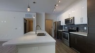 This single-floor residence showcases a thoughtfully designed interior with a contemporary aesthetic. At the heart of the space is an open-concept kitchen that immediately draws attention with its expansive white quartz island, which features an undermount sink and built-in electrical outlets — ideal for both meal preparation and casual dining. The kitchen is outfitted with a full suite of stainless steel appliances, including a French door refrigerator, electric range, and built-in microwave, all set against a backdrop of crisp white cabinetry and a classic gray subway tile backsplash. Track lighting runs along the ceiling, complementing the elegant pendant lights suspended above the island to create a warm, well-lit atmosphere. The flooring throughout is a rich, wood-look material that adds warmth and continuity to the space. A dedicated laundry area is conveniently tucked nearby, adding to the home's functional layout. Hallways connect to additional rooms, hinting at a well-organized floor plan designed for comfortable everyday living. The overall ambiance is clean, modern, and inviting, with a neutral color palette that lends itself to a variety of interior styles.