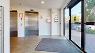 Located at 733 Goldstream Ave in Langford, BC, this building entrance lobby offers a clean, modern, and functional design. The space is finished with light wood-look luxury vinyl plank flooring throughout, complemented by crisp white walls and recessed ceiling lighting that create a bright, welcoming atmosphere. Large floor-to-ceiling glass entry doors flood the lobby with natural light and provide clear sightlines to the exterior streetscape and landscaping beyond.

A stainless steel elevator is prominently positioned within the lobby, providing accessible vertical access to upper floors. Adjacent to the elevator, a wall-mounted intercom and security panel allow for controlled building entry. A dedicated bank of individual resident mailboxes lines one wall, ensuring convenient mail retrieval for occupants.

The lobby also features a clearly marked Control Room door, along with additional utility access points and a stairwell door for fire egress. Safety signage, a fire alarm pull station, and a security camera are all present, reflecting the building's commitment to resident safety. A welcome mat at the main entrance and a sleek stainless steel waste bin near the entry doors add practical, everyday-use touches. The overall design is contemporary and efficiently organized, providing a tidy and functional first impression for residents and visitors alike.