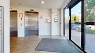 Located at 733 Goldstream Ave in Langford, BC, this building entrance lobby offers a clean, modern, and functional design. The space is finished with light wood-look luxury vinyl plank flooring throughout, complemented by crisp white walls and recessed ceiling lighting that create a bright, welcoming atmosphere. Large floor-to-ceiling glass entry doors flood the lobby with natural light and provide clear sightlines to the exterior streetscape and landscaping beyond.

A stainless steel elevator is prominently positioned within the lobby, providing accessible vertical access to upper floors. Adjacent to the elevator, a wall-mounted intercom and security panel allow for controlled building entry. A dedicated bank of individual resident mailboxes lines one wall, ensuring convenient mail retrieval for occupants.

The lobby also features a clearly marked Control Room door, along with additional utility access points and a stairwell door for fire egress. Safety signage, a fire alarm pull station, and a security camera are all present, reflecting the building's commitment to resident safety. A welcome mat at the main entrance and a sleek stainless steel waste bin near the entry doors add practical, everyday-use touches. The overall design is contemporary and efficiently organized, providing a tidy and functional first impression for residents and visitors alike.