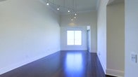 This single-floor space makes an immediate impression with its dramatically high ceilings that amplify the sense of openness throughout. The main living area is anchored by deep, richly toned hardwood floors that have been polished to a mirror-like finish, reflecting natural light and lending warmth and sophistication to the room. Crisp white walls enhance the airy, clean aesthetic, while a contemporary multi-pendant chandelier suspended from the ceiling serves as a refined focal point, casting soft, layered illumination across the space. A large window draws in abundant natural light, brightening the interior and offering views of the surrounding skyline or cityscape beyond. The architectural detailing is clean and precise, featuring classic white baseboards and a tray ceiling that adds subtle depth and dimension to the room's upper boundaries. The overall layout is open and unobstructed, providing exceptional flexibility for a variety of uses. The interplay of the gleaming floors, elevated ceiling height, and plentiful natural light creates an ambiance that is simultaneously elegant and inviting, suited for both functional living and refined entertaining.
