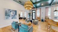 This thoughtfully designed single-floor space offers a welcoming and sophisticated environment spread across a generously proportioned open-plan layout. The interior is immediately distinguished by its dramatic teal-painted exposed beam ceilings, which contrast beautifully against crisp white walls and light hardwood flooring throughout.

At the heart of the space, a striking oversized woven rattan pendant chandelier anchors the main lounge area, where plush teal and gray upholstered armchairs are arranged around warm-toned round coffee tables atop layered area rugs. Large floor-to-ceiling windows dressed in sheer linen curtains flood the interior with abundant natural light and offer views of the surrounding exterior.

A prominent central island with a granite countertop and teal cabinetry serves as a functional focal point, illuminated by sleek pendant lights suspended from the beamed ceiling above. Bar-height seating lines the island, creating a casual gathering spot. Adjacent to this is a media lounge with a wall-mounted flat-screen television, shiplap wall paneling, and additional upholstered seating.

A separate dining nook features a marble-top round table with velvet chairs, bathed in natural light through tall windows. Curated artwork, including large-scale watercolor paintings and abstract canvases, is displayed throughout, lending the space a refined, gallery-like quality.